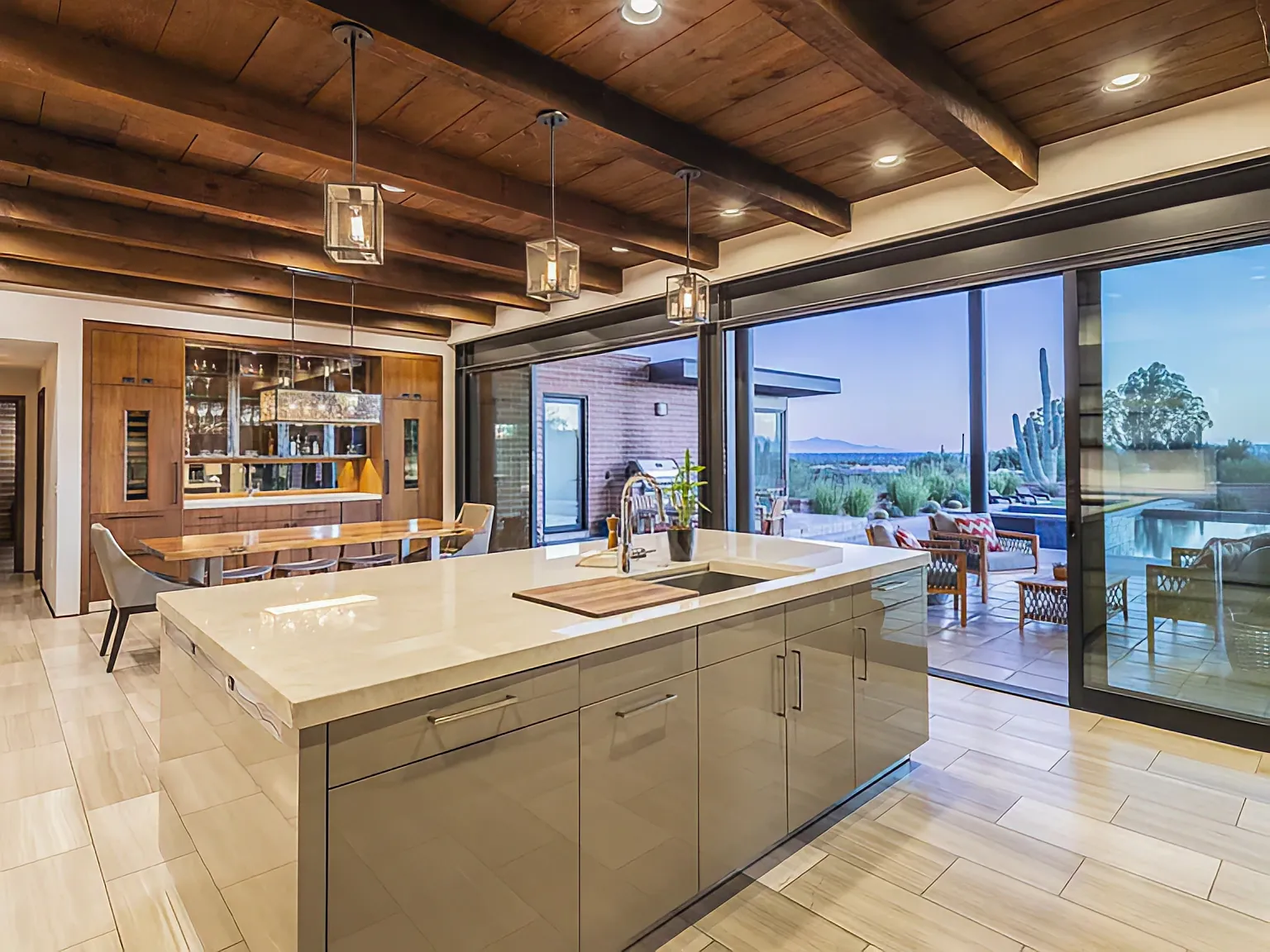 Kitchen and great room at Camino Antonio at dusk, Douglas Fir ceiling beams above stacking glass doors opened to the pool terrace and the desert beyond